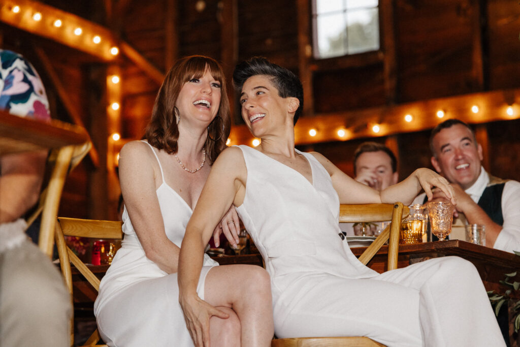 Two brides laughing joyfully together during an intimate wedding reception with warm string lights, candid documentary photography by Honey & Bloom