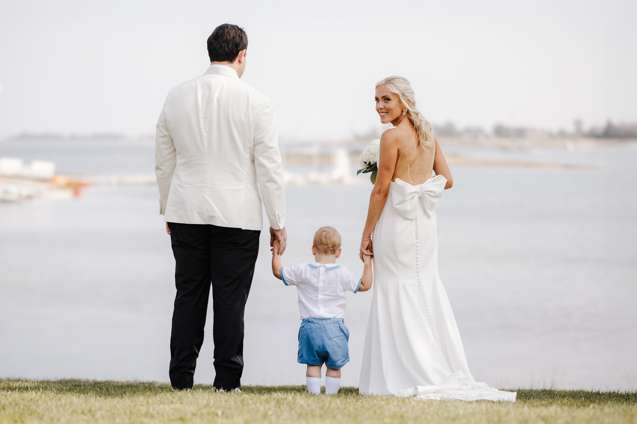 Newlyweds holding hands with a young child beside the water on their wedding day.