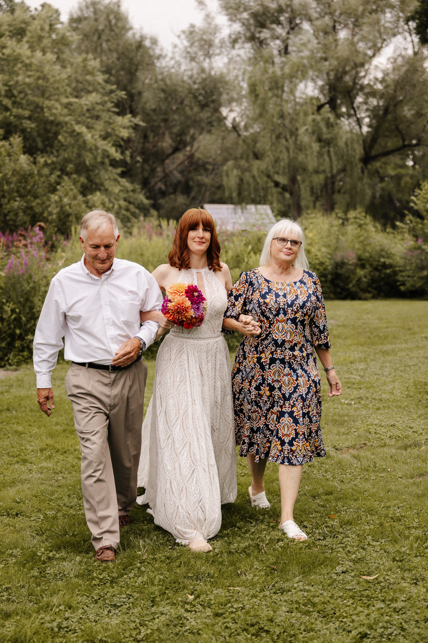 Bride walking arm in arm with two family members toward her outdoor wedding ceremony.