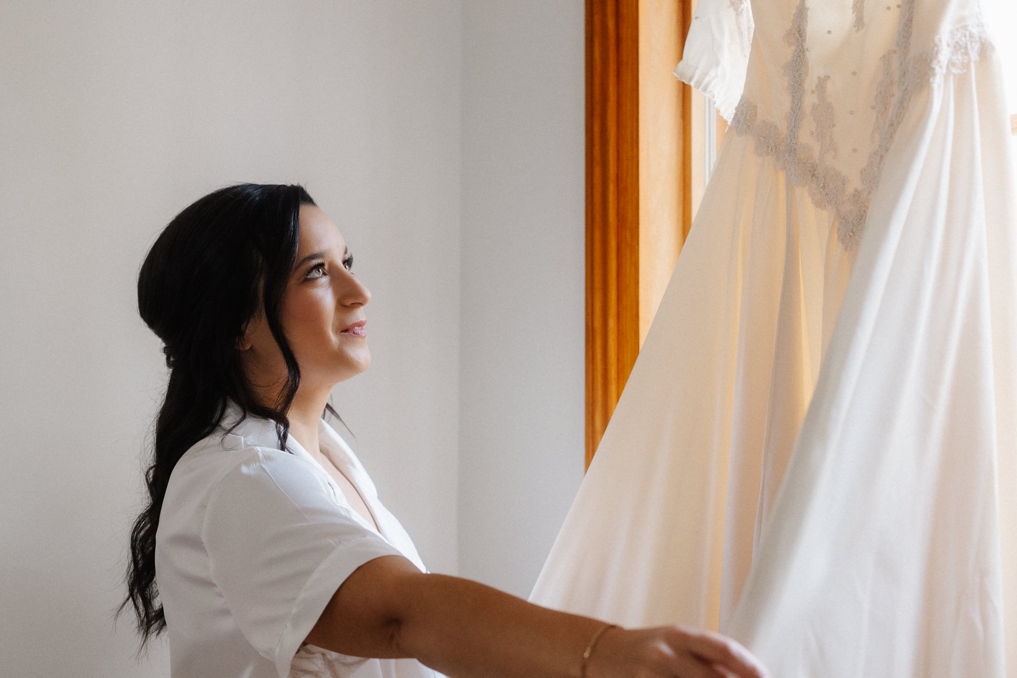 Bride reaching for her wedding dress hanging in window light on the wedding morning, Honey and Bloom Photography